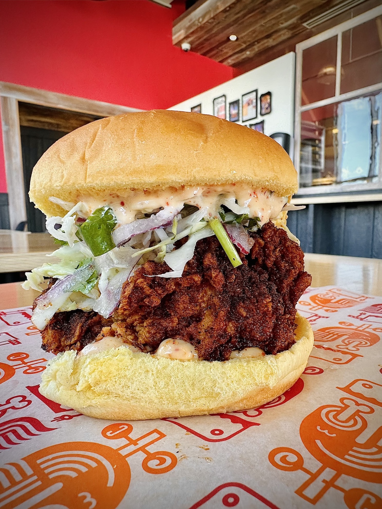 Crispy fried chicken sandwich featuring hand-breaded boneless thigh, topped with lime slaw and tajín mayo, served on a toasted potato bun, with a vibrant red restaurant backdrop.