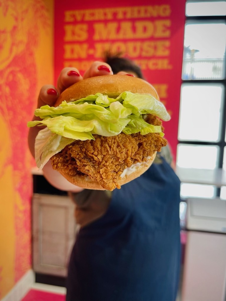 Crispy fried chicken sandwich with lettuce and mayo held up against a colorful background, showcasing Cluck N Cleaver's signature in-house preparation.