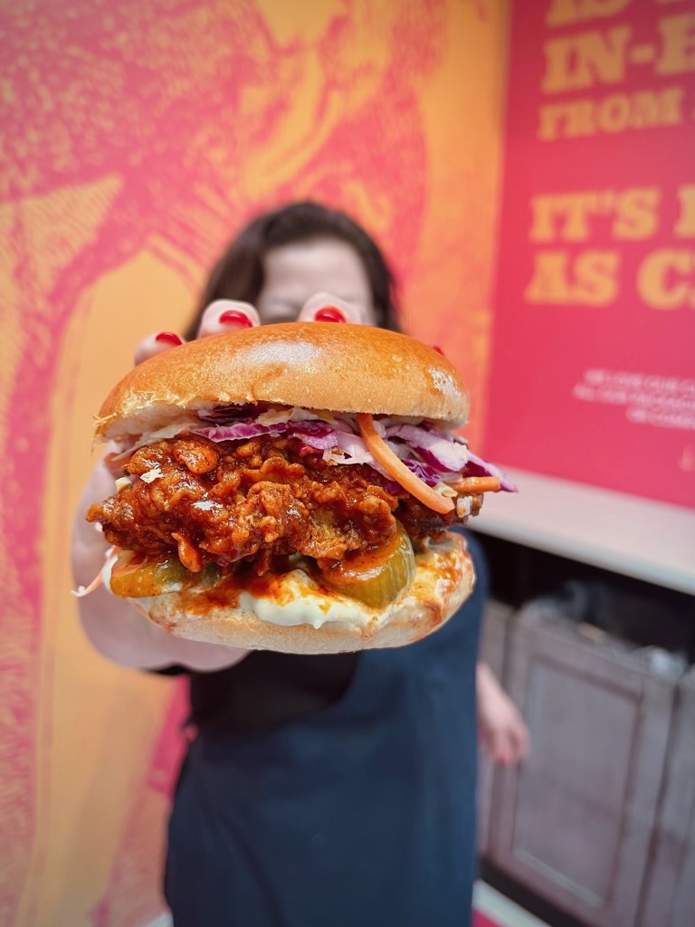 Crispy fried chicken sandwich featuring spicy sauce, crunchy slaw, and pickles on a toasted potato bun, held by a person against a vibrant background.