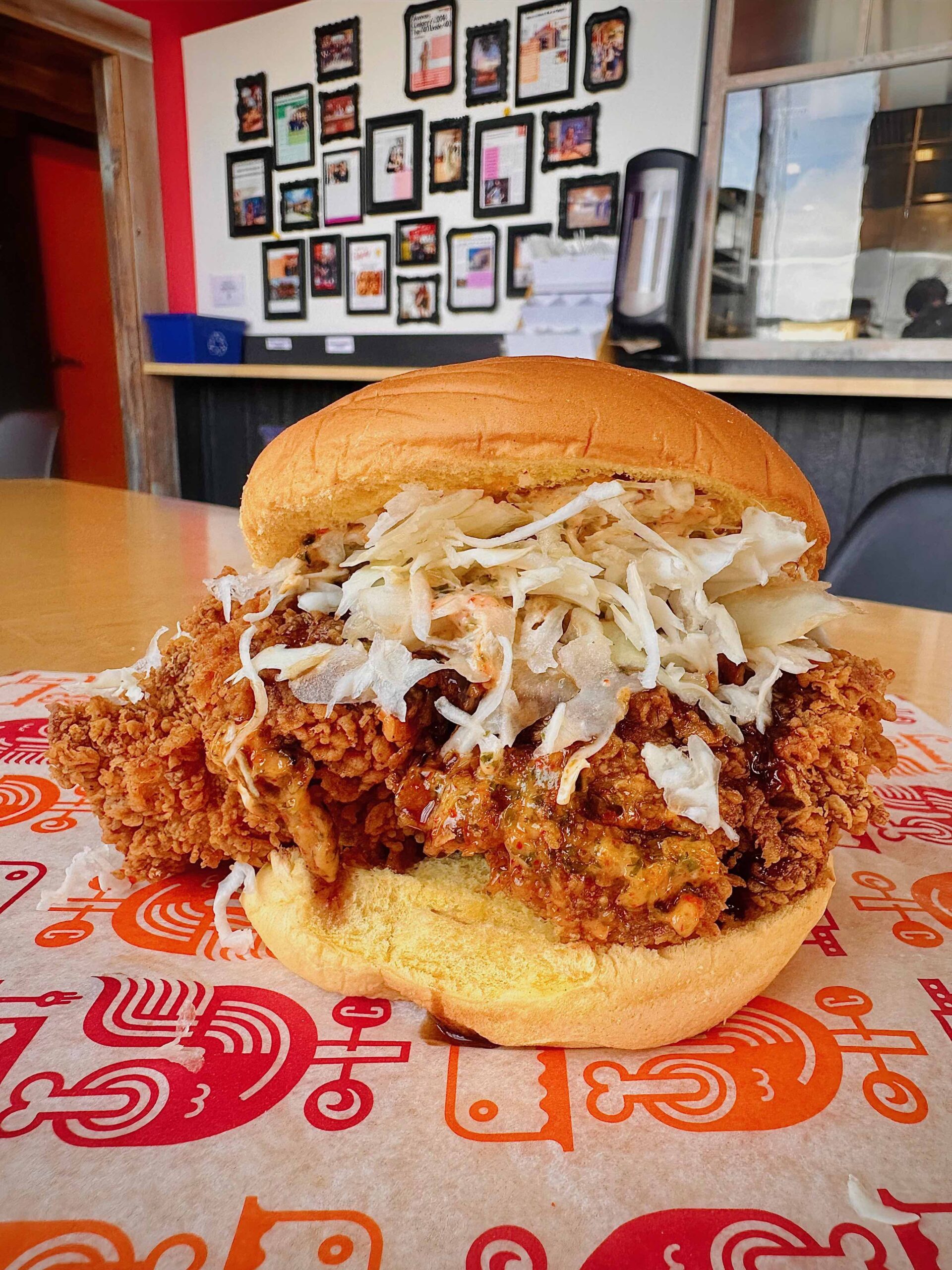 Crispy fried chicken sandwich featuring juicy boneless thigh, topped with shredded cabbage and house-made sauces, served on a toasted potato bun, with a vibrant restaurant backdrop.