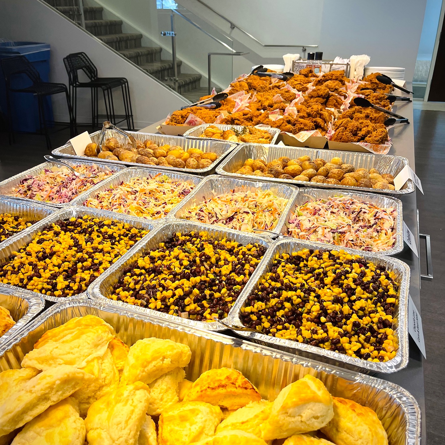 Fried chicken catering spread featuring crispy fried chicken pieces, assorted side dishes including coleslaw, corn salad, and biscuits, arranged for a corporate event or gathering.