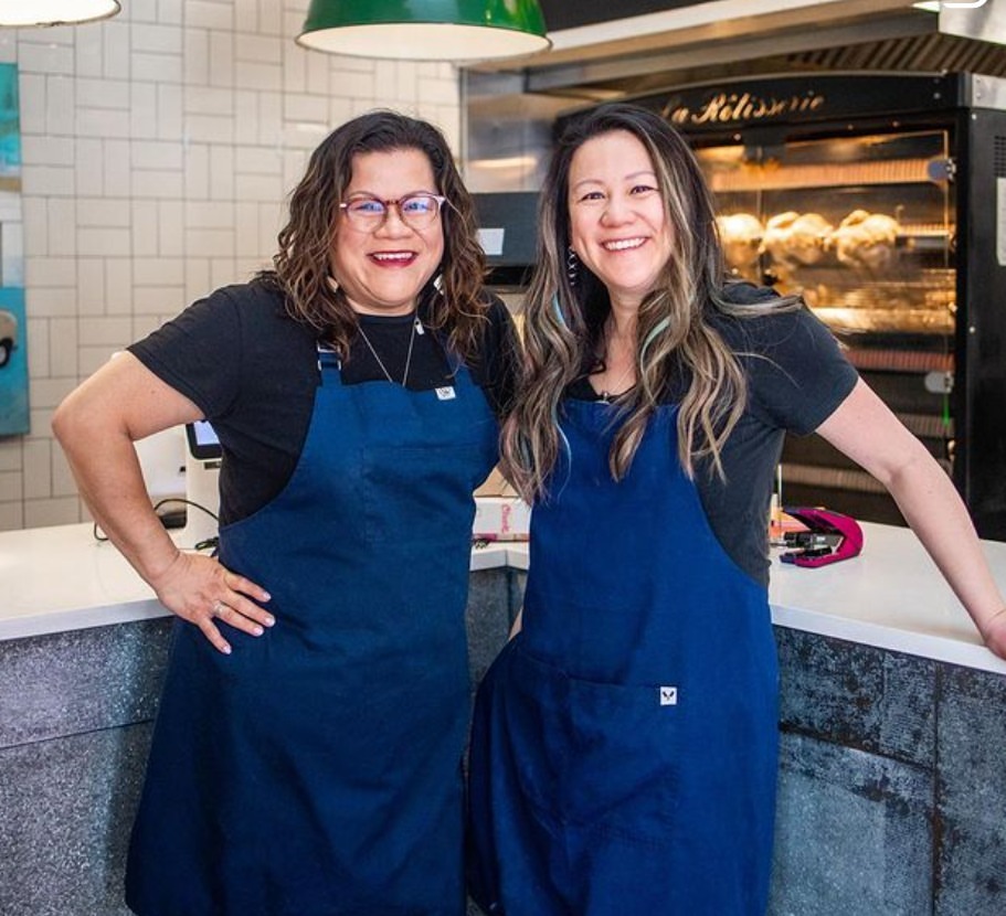 Chef Nicole Gomes and Francine Gomes, co-owners of Cluck N Cleaver, smiling in blue aprons at their Calgary restaurant, showcasing their commitment to quality and family-run operations in the fried chicken industry.